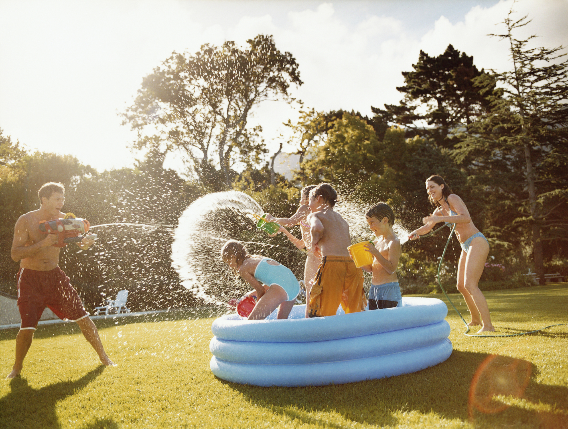 Father Aims A Water Gun At Children Throwing Water In A Paddling Pool