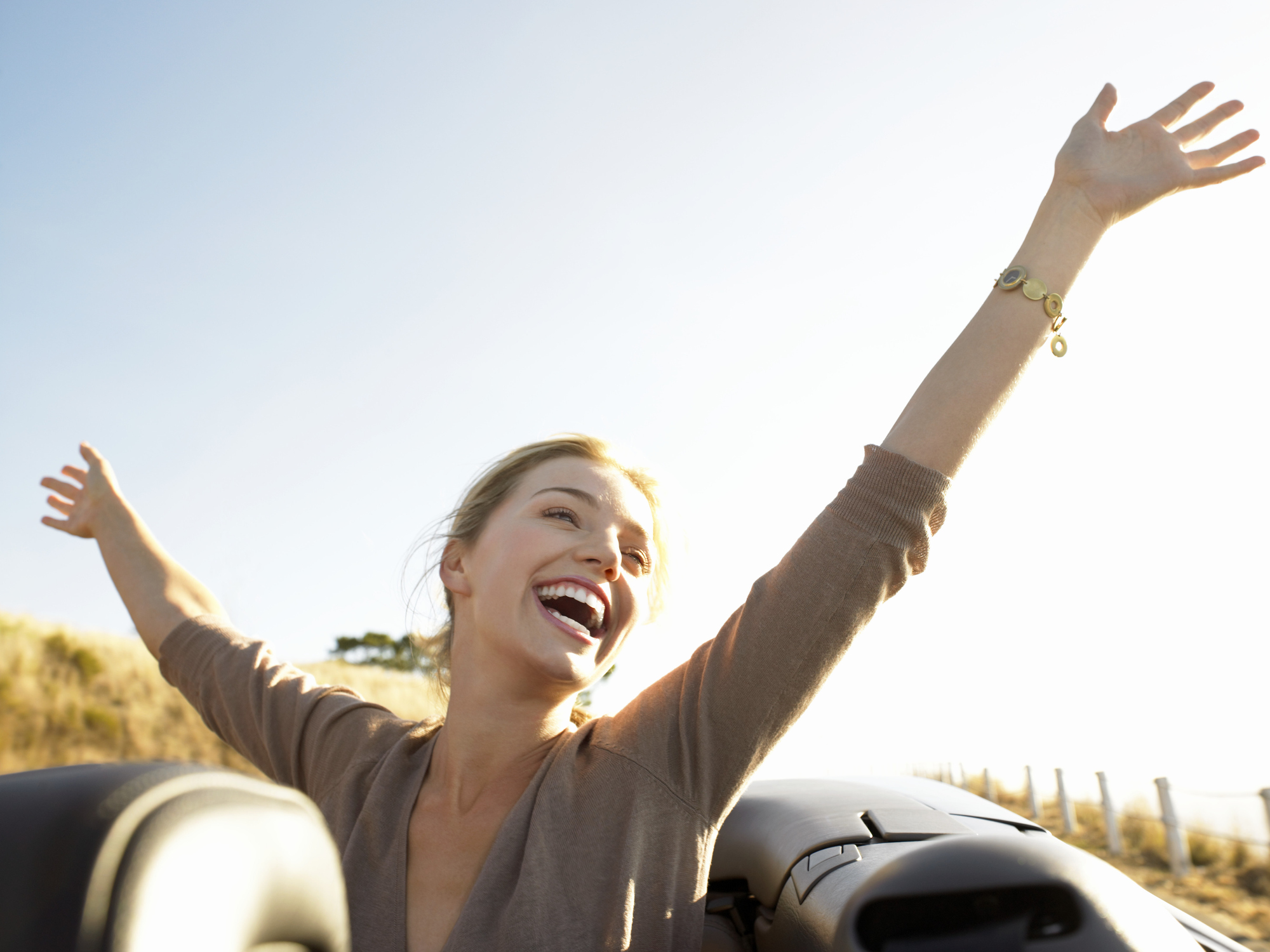 Young Woman Sits In The Back Of A Convertible, Her Arms In The Air, Laughing With Joy